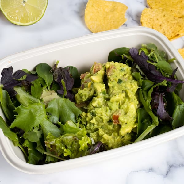 Guacamole and mixed greens in a white container, with tortilla chips and lime on marble.